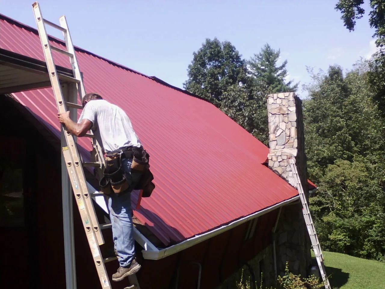 Roofer on a ladder beside a red metal roof and stone chimney — Soffit & Fascia Repair by Roofing Brothers in Orland Park